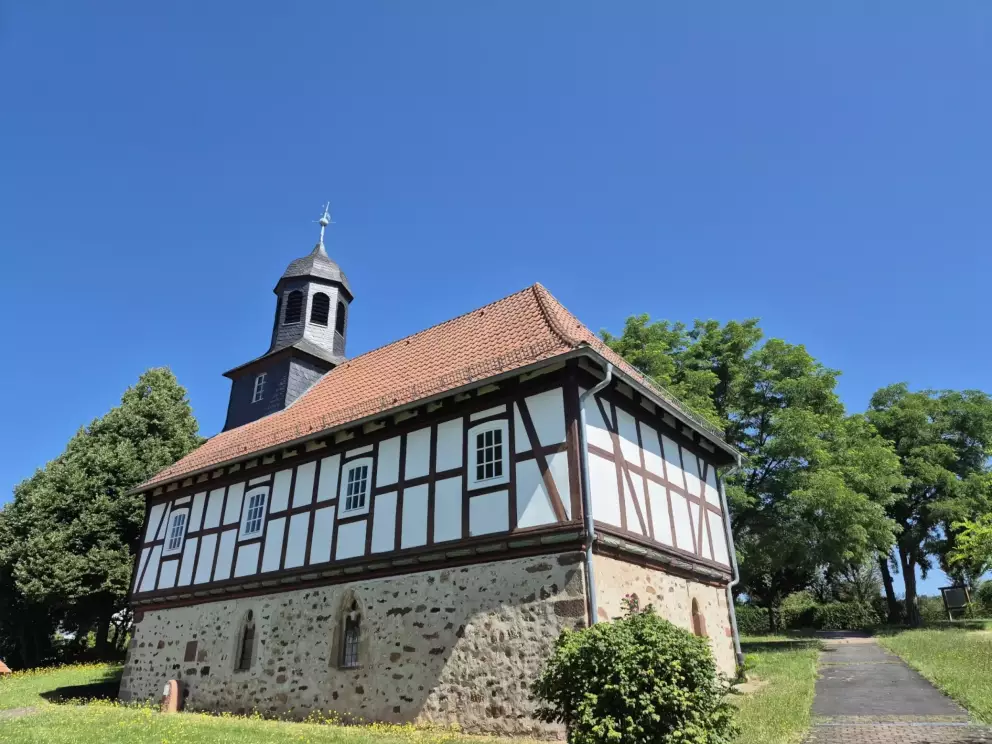 Fachwerkhaus mit 4 Fenstern, einem roten Dach und einem Glockenturm, im Hintergrund sind eine Wiese und Bäume