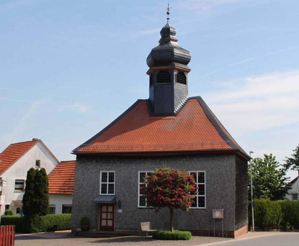 Kleine graue Kirche mit einem roten Dach, drei Fenstern und einem runder Baum