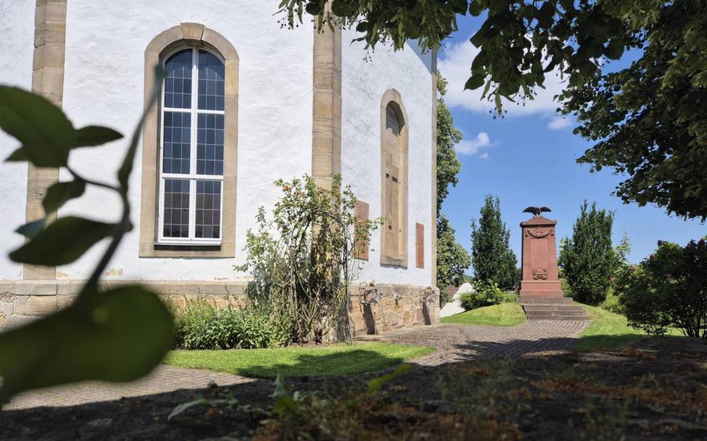 Ausschnitt einer weißen Kirche mit Rundbogenfenstern, im Hintergrund ein Denkmal
