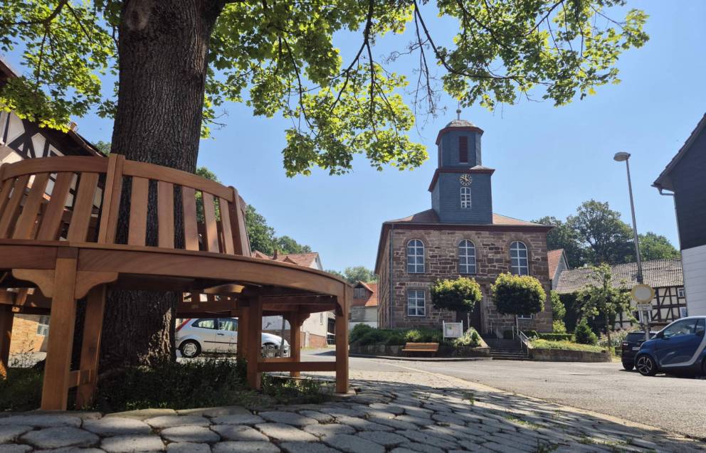 Runde Holzbank um einen Baum, dahinter ist eine kleine Backsteinkirche mit Rundbögenfenstern und einem Kirchturm