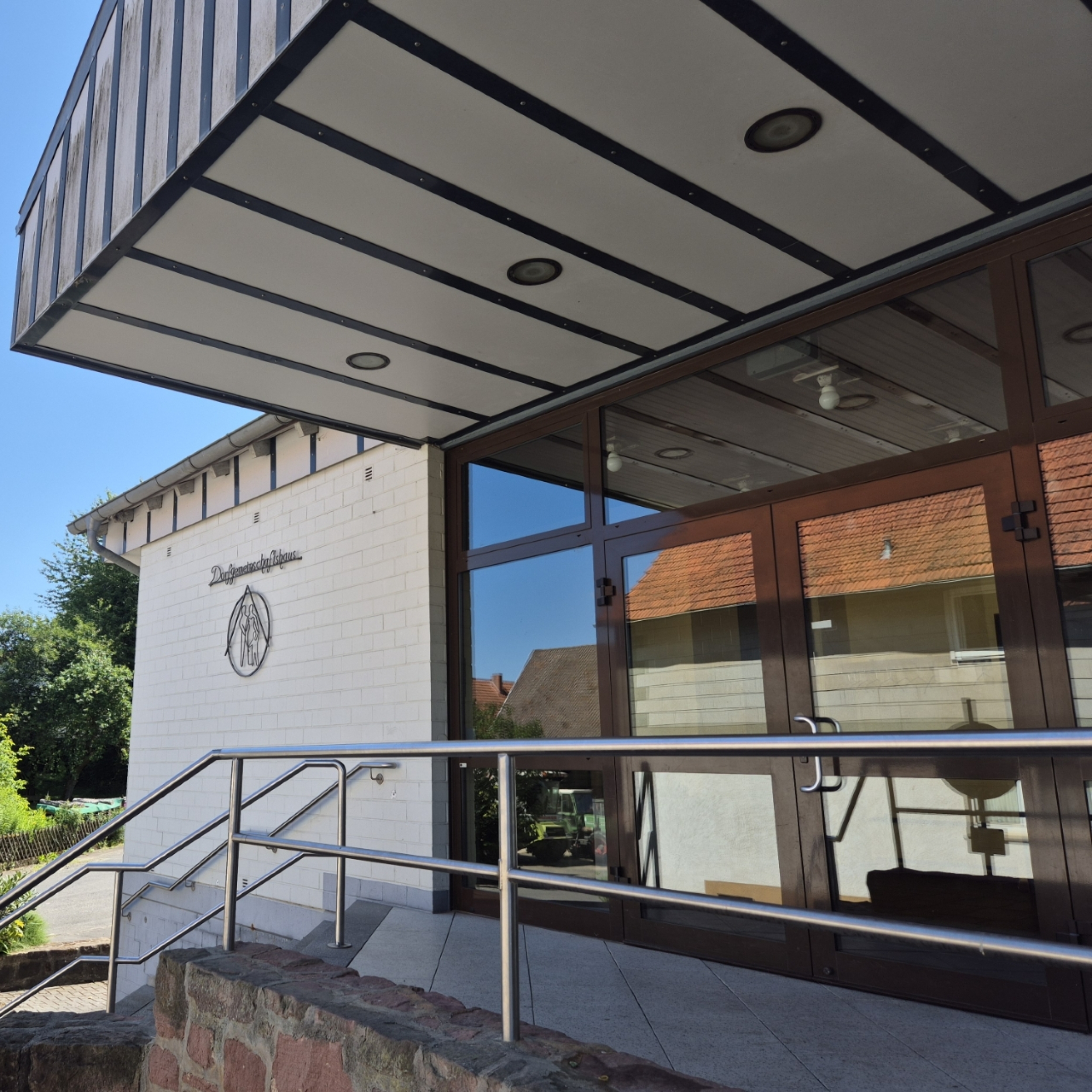 Eingang des Gemeinschaftshauses Ascherode, davor eine Treppe mit einem Edelstahlgeländer einer breiten vierteiligen Glaseingangstür und einer Überdachung