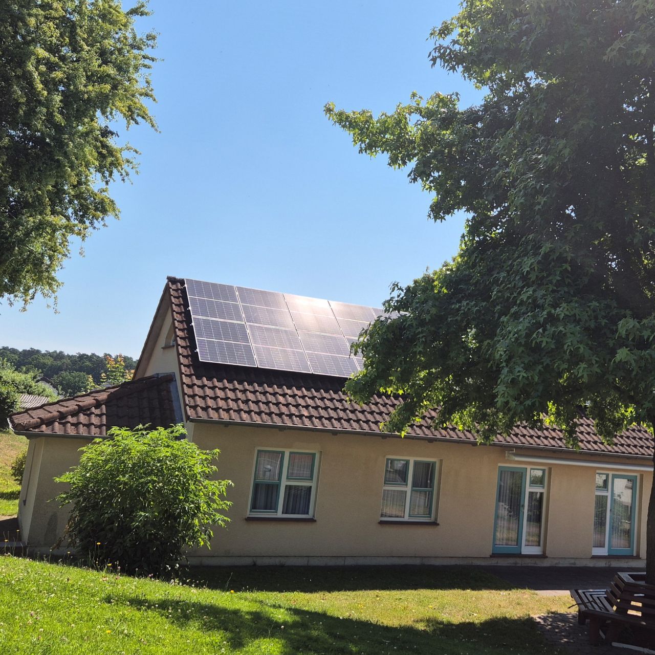 Aussenansicht Gemeinschaftshaus Floshain, einstöckiges Gebäude mit Fenstern und einer Solaranlage auf dem Dach im Vordergrund eine runde Holzbank die um einen Baum gebaut ist
