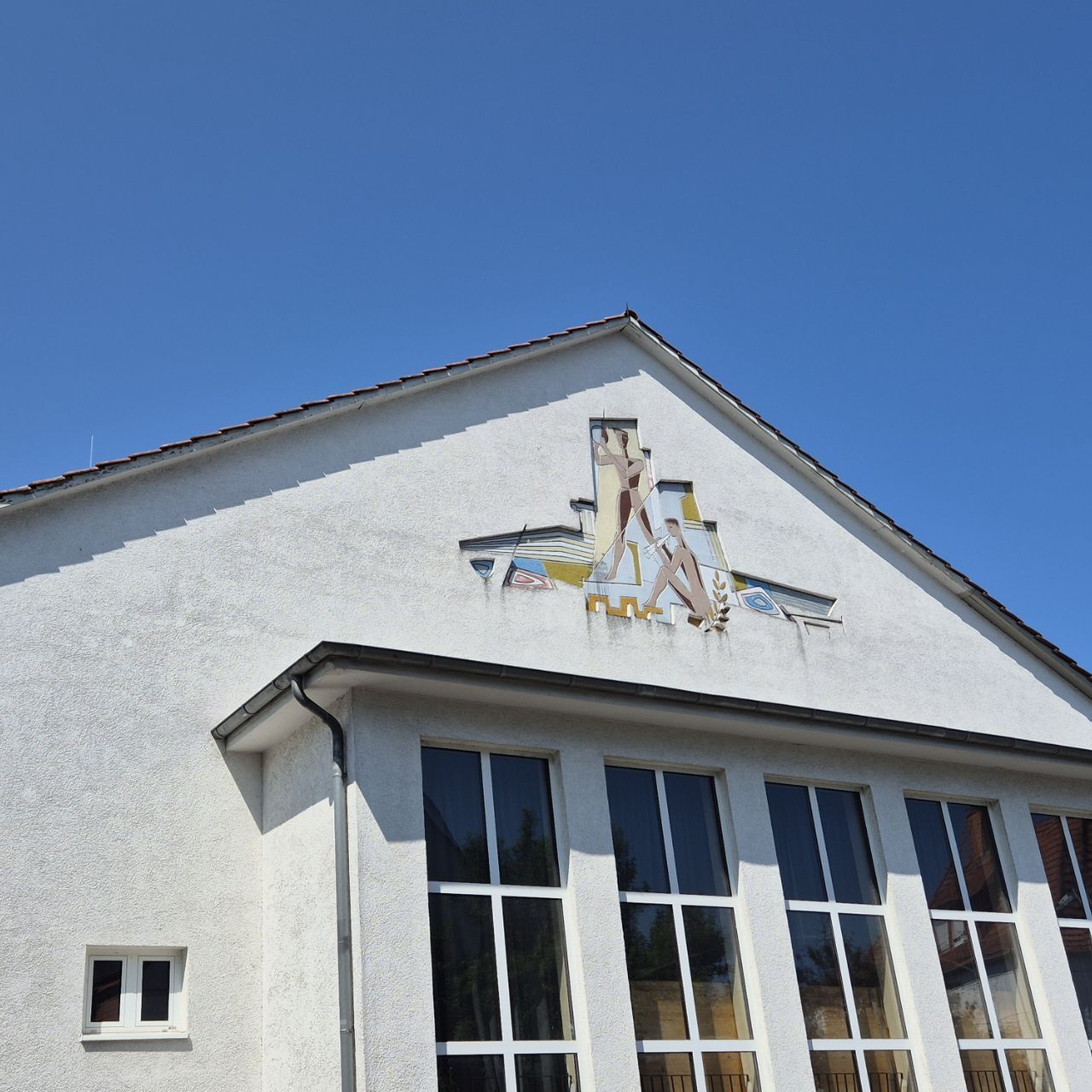 Aussenansicht Sport- und Kulturhalle Ziegenhain, weisses Gebäude von der Seite fotografiert, vier weiße Fenster darüber eine Malerei im Giebel 