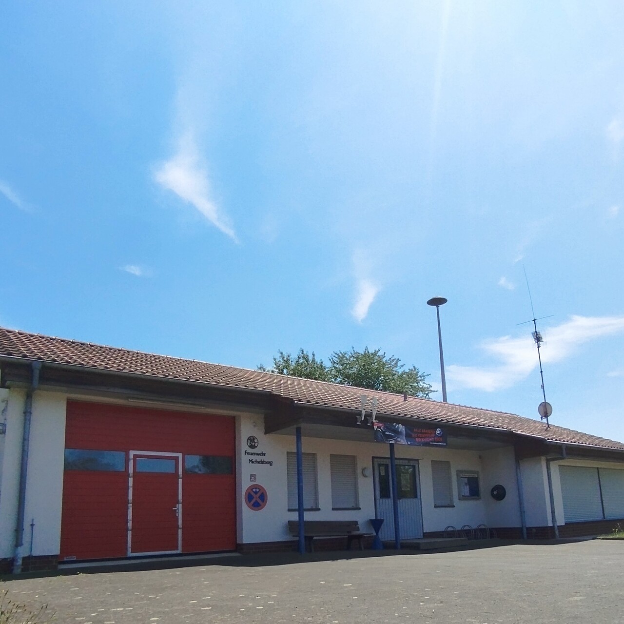 Gemeinschaftshaus Michelsberg von außen, weißes Gebäude mit einem roten Dach, links neben dem Eingang ein rotes Tor für die Feuerwehr, rechts daneben zwei große Fenster mit heruntergelassenen Rolläden